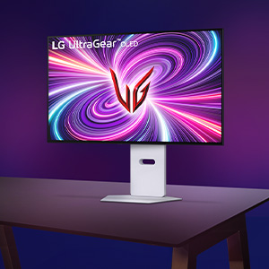 보라색 그라데이션 배경의 어두운 책상 위에 LG 울트라기어 모니터가 놓여 있습니다. 모니터 화면에는 분홍색과 보라색이 섞인 화려한 소용돌이 패턴 중앙에 빨간색 'UG' 로고가 배치되어 있으며, 화면 왼쪽 상단에는 'LG UltraGear OLED'라는 문구가 적혀 있습니다.