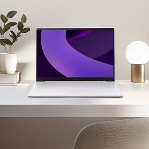 깨끗하고 밝은 화이트 톤의 책상 위에 화이트 색상의 노트북이 정면을 향해 열려 있습니다. 노트북 화면에는 보라색의 부드러운 곡선이 겹쳐진 추상적인 배경화면이 나타나 있습니다. 책상 왼쪽에는 작은 화분이, 오른쪽에는 둥근 형태의 은은한 조명이 배치되어 있어 깔끔하고 차분한 작업 환경을 보여줍니다.